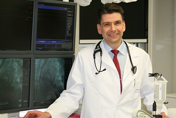 Ein lächelnder Arzt im weißen Kittel mit Stethoskop steht in einem klinischen Untersuchungsraum vor mehreren Monitoren mit medizinischen Bildern. Er wirkt bereit für eine Untersuchung oder Beratung.