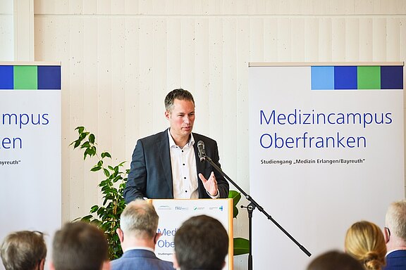 Ein Redner spricht am Pult vor Publikum bei einer Veranstaltung des „Medizincampus Oberfranken“. Aufstellbanner zeigen den Titel und den Studiengang „Medizin Erlangen/Bayreuth“. Mikrofon und Rednerpult im Vordergrund; Vortragssituation in heller, schlichter Umgebung.