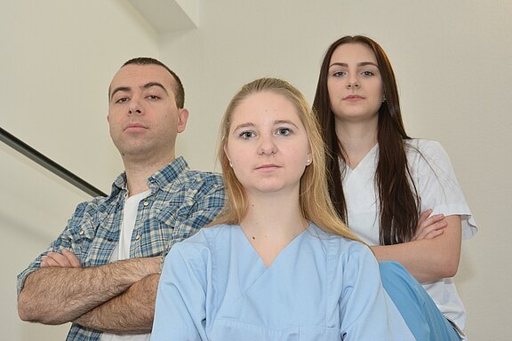 Drei Personen stehen in einem Raum. Die Frau in der Mitte trägt ein hellblaues Oberteil, sie hat lange, blonde Haare und schaut ernst. Rechts steht eine Frau in einem weißen Oberteil mit langen, dunklen Haaren. Links steht ein Mann in einem karierten Hemd, ebenfalls mit ernstem Ausdruck.