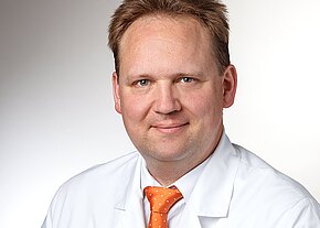 Dr. med. Christoph Braig Porträt eines mittelalten Mannes in weißem Kittel mit orangefarbener Punktkrawatte, leicht lächelnd, frontal in die Kamera blickend vor neutralem Hintergrund.