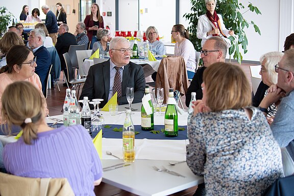 Mehrere Erwachsene sitzen in einem hellen Saal an runden Tischen, trinken Wasser oder Saft und unterhalten sich. Im Vordergrund spricht ein älterer Mann im Anzug mit seinen Tischnachbarn. Insgesamt wirkt es wie ein zwangloses Treffen oder Empfang.