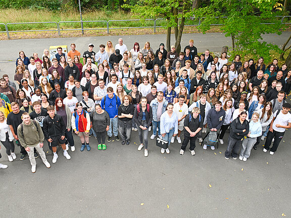 Eine große Gruppe von Menschen steht im Freien, auf einem gepflasterten Platz. Die Personen sind vielfältig gekleidet und lächeln in die Kamera. Bäume und Wiese sind im Hintergrund sichtbar. Die Anordnung der Gruppe vermittelt ein Gefühl von Gemeinschaft und Zusammengehörigkeit.