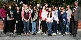 Eine Gruppe junger Frauen steht im Freien, umgeben von Bäumen. Sie sind in legerer Kleidung gekleidet und lächeln in die Kamera. Einige Mitglieder der Gruppe tragen Namensschilder. Die Stimmung wirkt freundlich und kollegial.