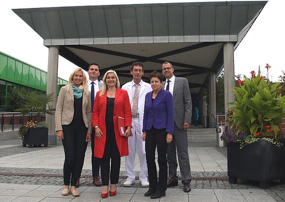 Sechs Erwachsene posieren vor dem überdachten Eingangsbereich eines Gebäudes. Fünf tragen Businesskleidung, eine Person einen weißen Arztkittel. Sie stehen nebeneinander, schauen in die Kamera und lächeln. Der Fokus liegt auf der Gruppe.