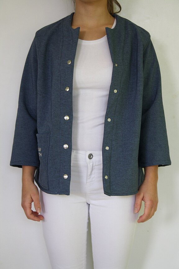 Person trägt offene, dunkelgraublaue Jacke mit Druckknöpfen und 3/4-Ärmeln über weißem T‑Shirt und weißer Hose. Fokus auf der Jacke, vorne offen, schlichte, gerade Form.