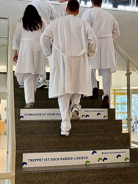 Mehrere Personen in weißen Kitteln steigen eine Treppe hinauf. Auf den Stufen stehen motivierende Sprüche wie „Treppe? Ist doch kardio-logisch“ und „Schnaufen ist keine Schande“. Die Szene wirbt für Bewegung und die gesundheitlichen Vorteile des Treppensteigens.