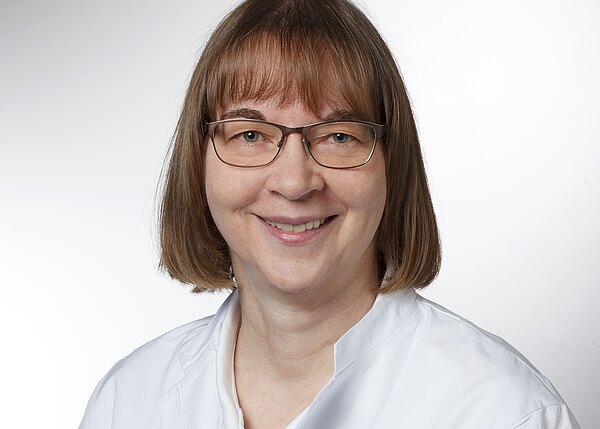 Porträt einer lächelnden Frau mit schulterlangem braunen Haar und Brille, in weißem medizinischen Kittel, vor hellem neutralem Hintergrund.