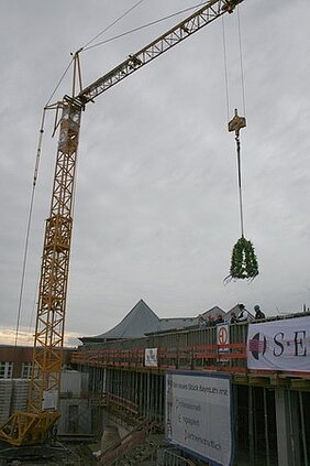 Ein Kran hebt einen Kranz über eine Baustelle, während Arbeiter auf dem Gerüst stehen. Im Hintergrund sind Teile eines Gebäudes und Werbebanner zu sehen. Der Himmel ist bewölkt und sorgt für eine düstere Atmosphäre.