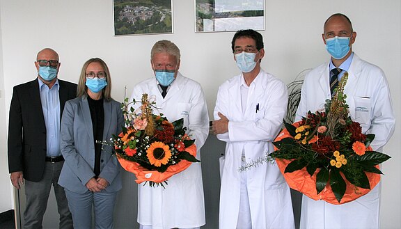 Fünf Personen stehen in einem Raum, alle tragen Mund-Nasen-Schutz. Vier Personen tragen weiße Laborkittel und halten große Blumensträuße. Eine Person trägt einen grauen Anzug. Im Hintergrund sind Bilder an der Wand zu sehen.