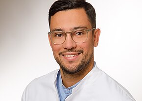 Dr. Maximilian Klumm Porträt eines lächelnden Mannes mit Brille in weißem Arztkittel vor hellem, neutralem Hintergrund.