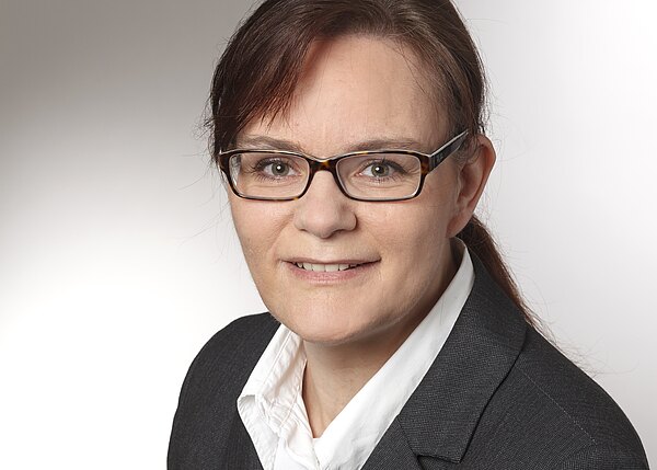 Professionelles Porträt: Frau mit Brille, braunen Haaren und Pferdeschwanz, in dunklem Blazer und weißer Bluse, lächelt leicht vor neutralem Hintergrund.