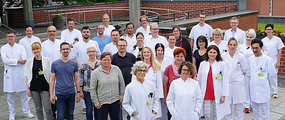 Gruppenfoto eines großen Krankenhaus-Teams: Ärztinnen, Ärzte und Pflegekräfte, viele in weißen Kitteln, einige in Freizeitkleidung, stehen draußen auf dem Klinikgelände und schauen in die Kamera. Fokus: gemeinsames medizinisches Team.