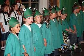 Eine Gruppe von Kindern steht in einer Reihe und trägt grüne Gewänder sowie Froschkappen. Sie wirken begeistert und singen zusammen. Im Hintergrund sind weitere Kinder und eine Person im Rollstuhl zu sehen.