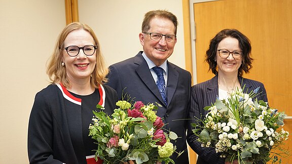 Drei Personen stehen zusammen und halten große Blumensträuße. Sie lächeln und tragen formelle Kleidung. Der Hintergrund ist neutral gehalten. Es scheint eine feierliche Atmosphäre zu herrschen.