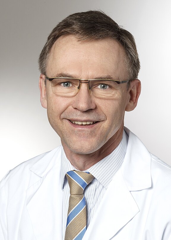 Porträt eines lächelnden Mannes mittleren Alters mit Brille, weißem Arztkittel, Hemd und gestreifter Krawatte vor neutralem Hintergrund.