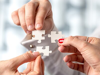 Mehrere Hände halten vier weiße Puzzleteile und setzen sie zusammen. Nahaufnahme in neutraler Umgebung. Symbol für Zusammenarbeit, Vernetzung und das Finden einer passenden Lösung.