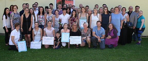Große Gruppe lächelnder Erwachsener posiert im Freien vor einer hellen Wand. In der ersten Reihe halten mehrere Personen Urkunden und große Schecks. Die Szene wirkt wie eine Preisverleihung oder Abschlussfeier im Pflege- oder Bildungsbereich.