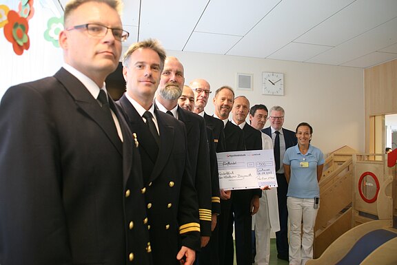 Eine Gruppe uniformierter Männer überreicht in einer Krankenhaus-Kinderstation einen großen Spendenscheck. Neben ihnen stehen Ärztinnen und Ärzte sowie Pflegepersonal, lächelnd. Der Schwerpunkt liegt auf der Spendenübergabe.