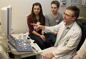 Ein Arzt im weißen Kittel erklärt etwas an einem Ultraschallgerät. Zwei Jugendliche, ein Mädchen und ein Junge, hören aufmerksam zu und schauen auf den Bildschirm. Der Arzt zeigt mit dem Finger auf eine Stelle auf dem Bildschirm.
