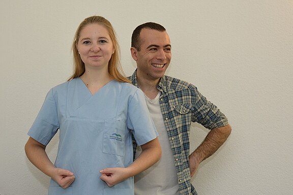 Eine junge Frau in einem blauen Krankenhauskleid mit verschränkten Armen steht vorne. Hinter ihr lächelt ein Mann in einem karierten Hemd und hat die Hände in den Hüften. Beide stehen vor einer hellen Wand.