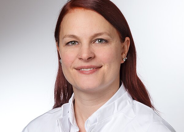 Porträtfoto: Eine lächelnde Person mit rötlich-braunen, schulterlangen Haaren trägt ein weißes Oberteil mit Kragen. Heller, neutraler Hintergrund; der Fokus liegt auf dem Gesicht und dem freundlichen Ausdruck.