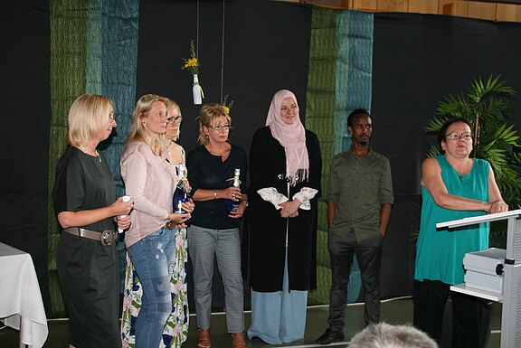 Auf einer Bühne steht eine Gruppe Erwachsener nebeneinander; einige halten Blumen und Geschenkflaschen. Rechts spricht eine Frau am Redepult. Die Szene wirkt wie eine Ehrung oder Verabschiedung.