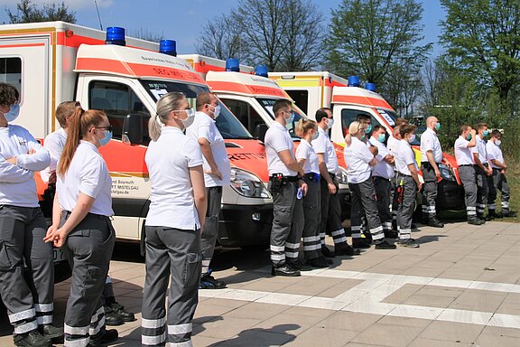 Eine Gruppe von Rettungssanitätern in einheitlicher Kleidung steht auf einem Platz vor mehreren Rettungswagen. Alle tragen Mund-Nasen-Schutz. Die Sanitäter schauen in eine Richtung, während sich Bäume im Hintergrund abzeichnen.