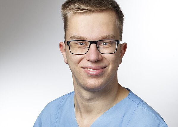 Nahaufnahme eines jungen Mannes mit Brille, kurz geschnittenen Haaren und freundlichem Lächeln. Er trägt ein hellblaues Oberteil, ähnlich medizinischer OP-Kleidung. Neutraler, heller Hintergrund.