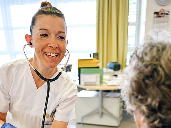 Pflegekraft in weißer Kleidung und blauen Handschuhen misst mit Stethoskop und Blutdruckmanschette den Blutdruck einer älteren Person am Arm. Freundliche, beruhigende Situation in einer Praxis.