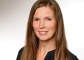 Porträtfoto einer lächelnden Person mit langen braunen Haaren, schwarzem Oberteil, vor hellem neutralem Hintergrund.