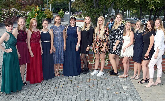 Gruppe junger Frauen steht nebeneinander und lächelt in die Kamera. Sie tragen überwiegend lange, elegante Kleider in verschiedenen Farben; eine trägt ein geblümtes kurzes Kleid mit weißen Turnschuhen. Aufnahme im Freien.