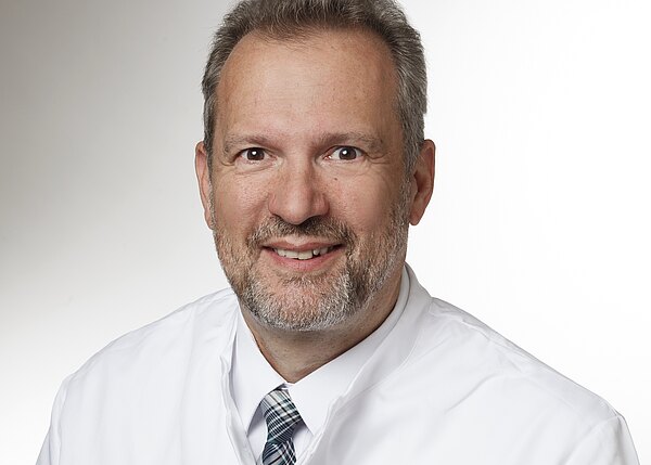 Porträt eines lächelnden Mannes mittleren Alters im weißen Arztkittel mit kariierter Krawatte, vor neutralem Hintergrund. Nahaufnahme, Kopf und Schultern, direkter Blick in die Kamera.