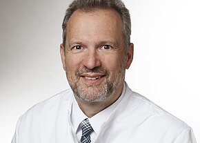 Porträt eines lächelnden Mannes mittleren Alters im weißen Arztkittel mit kariierter Krawatte, vor neutralem Hintergrund. Nahaufnahme, Kopf und Schultern, direkter Blick in die Kamera.