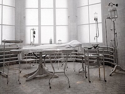 Historischer Operationssaal: leeres OP-Bett mit weißem Tuch, umgeben von rollbaren Metalltischen, Instrumenten und Infusionsständern. Heller, rundes Fensterband, gefliester Boden, nüchterne, sterile Atmosphäre. Keine Personen, nur alte medizinische Geräte.