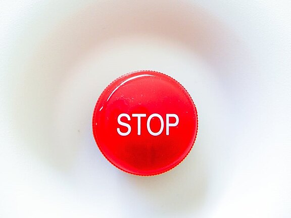 Zentral platzierter roter runder Knopf mit weißer Aufschrift STOP auf hellem, minimalistischem Hintergrund; wirkt wie ein Not-Aus-Schalter.