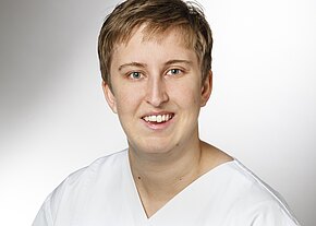 Station 3a/3b Porträt einer lächelnden Person mit kurzem Haar in weißem Kasack, frontal aufgenommen vor hellem, neutralem Hintergrund. Der Fokus liegt auf dem freundlichen Gesicht.