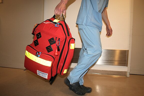 Medizinische Fachkraft in blauer OP-Kleidung geht durch einen Krankenhausflur und trägt eine große rote Notfalltasche mit reflektierenden Streifen.