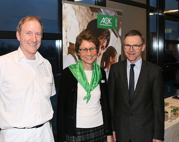Drei Personen stehen nebeneinander und lächeln bei einer AOK‑Veranstaltung. In der Mitte eine Frau mit grünem Schal, links ein Mann in weißer Kleidung, rechts ein Mann im dunklen Anzug. Hintergrund: Infostand mit Bannern.