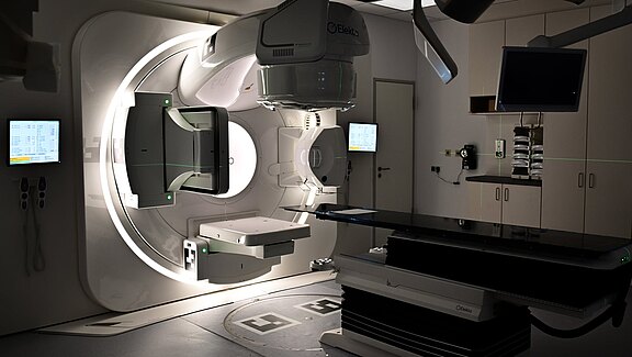 Ein modernes Bestrahlungszimmer mit einem großen, kreisförmigen Gerät in der Mitte, das für die Strahlentherapie genutzt wird. Rechts befindet sich ein Behandlungstisch. An der Wand hängen Bildschirme, die medizinische Daten anzeigen. Das Zimmer ist gut beleuchtet und wirkt klinisch.