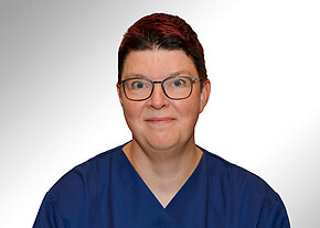 Stefanie Hopf Porträt einer Person mit kurzer, dunkelroter Frisur und Brille, die blaue medizinische Kleidung trägt, vor hellem, neutralem Hintergrund. Die Person blickt lächelnd in die Kamera.