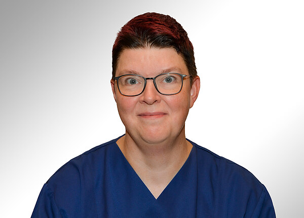 Porträt einer Person mit kurzer, dunkelroter Frisur und Brille, die blaue medizinische Kleidung trägt, vor hellem, neutralem Hintergrund. Die Person blickt lächelnd in die Kamera.