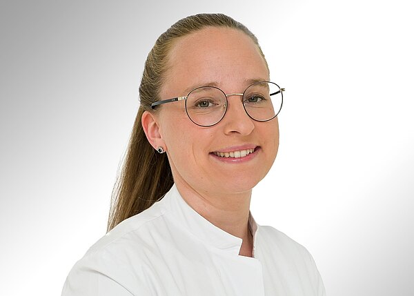 Porträt einer lächelnden Person mit runder Brille und zurückgebundenen Haaren, in weißem Kittel vor hellem Hintergrund, Blick zur Kamera.