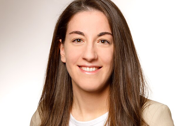 Portrait einer lächelnden Frau mit langen, braunen Haaren. Sie trägt ein weißes T-Shirt und einen hellen Blazer. Der Hintergrund ist neutral und hell, wodurch ihr Ausdruck freundlich und einladend wirkt.