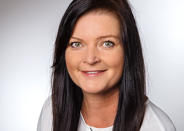 Porträtfoto: Frau mit langen dunklen Haaren und hellen Augen, freundlich lächelnd, trägt eine weiße Bluse. Neutraler Studiohintergrund, Kopf-und-Schultern-Aufnahme, Blick direkt in die Kamera.