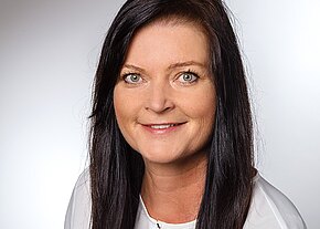 Porträtfoto: Frau mit langen dunklen Haaren und hellen Augen, freundlich lächelnd, trägt eine weiße Bluse. Neutraler Studiohintergrund, Kopf-und-Schultern-Aufnahme, Blick direkt in die Kamera.