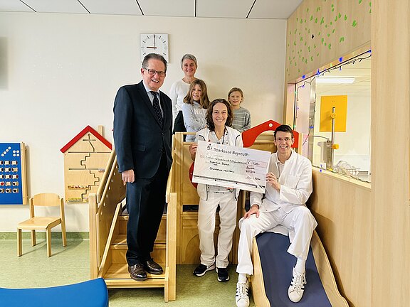 In einem hellen Kinderbereich einer Klinik posieren Mitarbeitende in weißer Kleidung mit einem großen Spendenscheck. Weitere Erwachsene und zwei Kinder stehen daneben und lächeln.