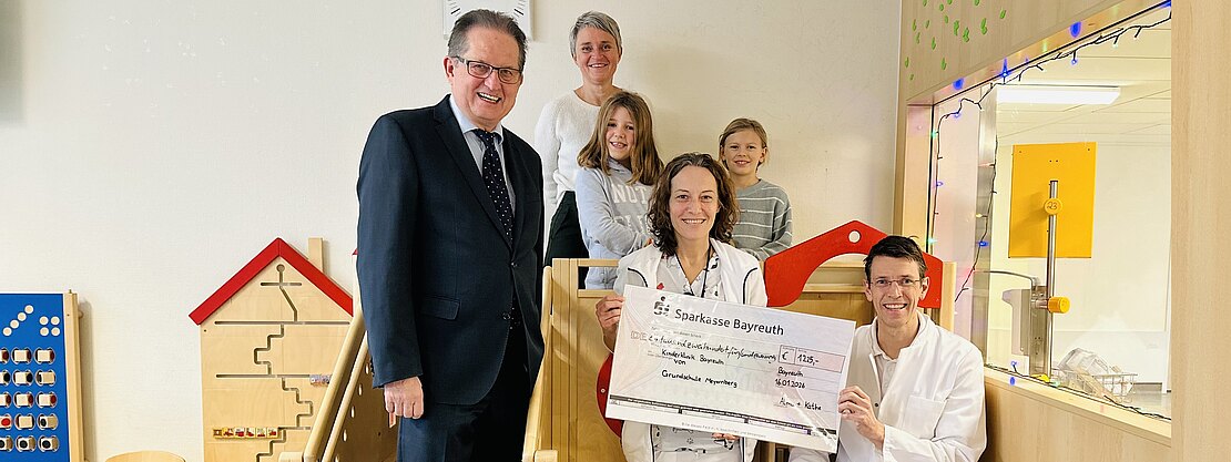 In einem hellen Kinderbereich einer Klinik posieren Mitarbeitende in weißer Kleidung mit einem großen Spendenscheck. Weitere Erwachsene und zwei Kinder stehen daneben und lächeln.