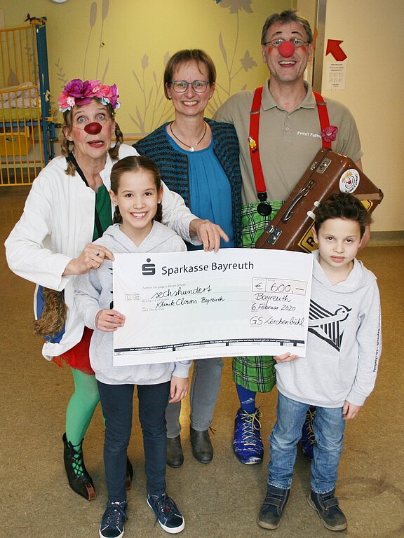 Gruppenfoto bei einer Spendenübergabe: Zwei Kinder halten einen großen Scheck über 600 Euro, daneben zwei verkleidete Klinikclowns und eine Frau. Der Scheck ist von der Sparkasse Bayreuth und geht an Klinikclowns. Fröhliche Stimmung, Innenraum.