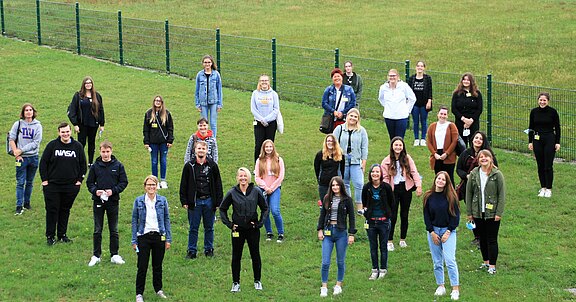 Eine Gruppe von 30 Personen steht auf einer grünen Wiese. Sie sind lächelnd angeordnet, einige tragen Jacken und Hoodies. Im Hintergrund ist ein Zaun sichtbar. Es ist eine freundliche, gesellige Stimmung.