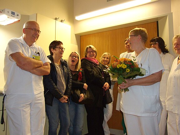 Person im Pflegekittel hält einen Blumenstrauß, umringt von lachenden Kolleginnen, Kollegen und Besuchern in einem Krankenhausflur; die Szene wirkt wie eine kleine Feier oder Dankesgeste.